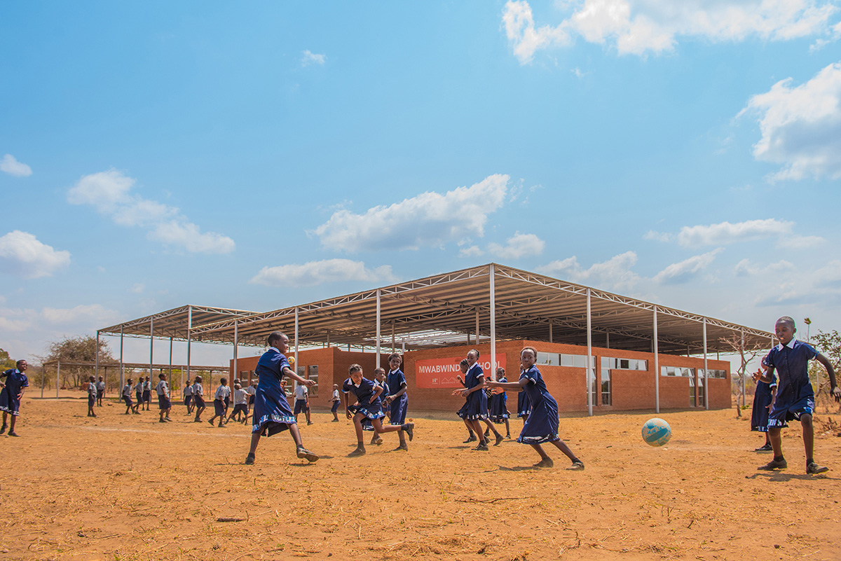非洲儿童在砖砌学校前踢足球，学校的招牌写着Mwabwindo